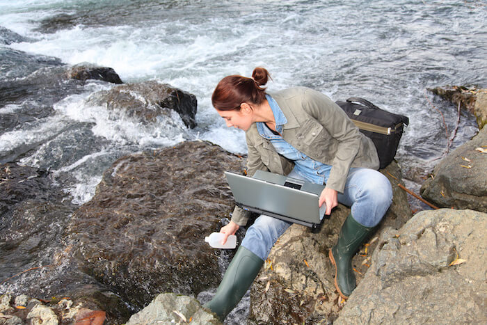 A scientist in wellington boots with her laptop sits on a rock and takes a water sample from a river