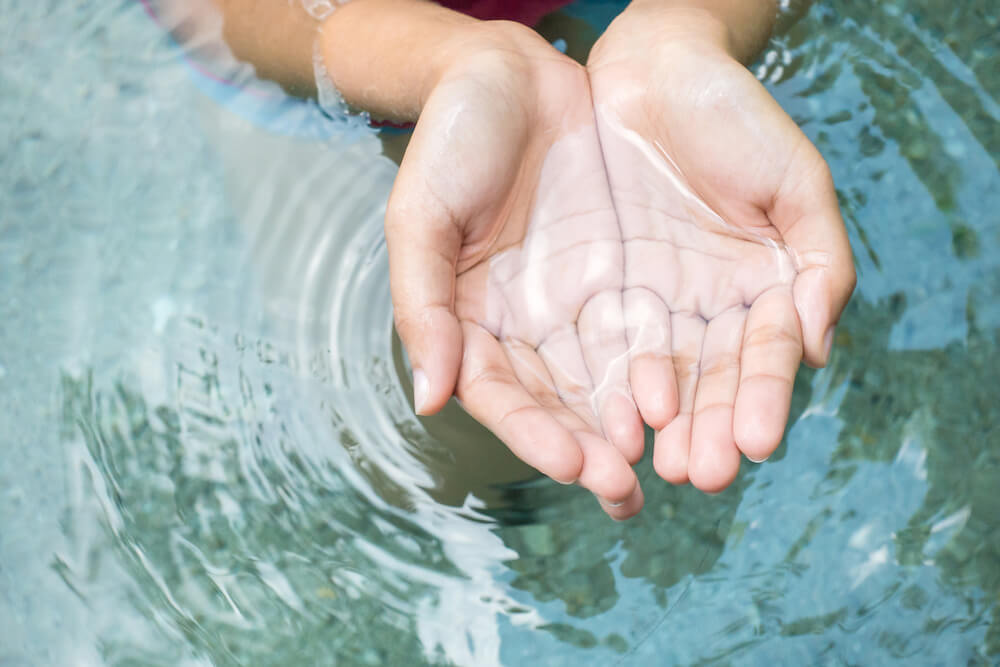 A person's hands are cupped in clear, natural water as if to drink from it.