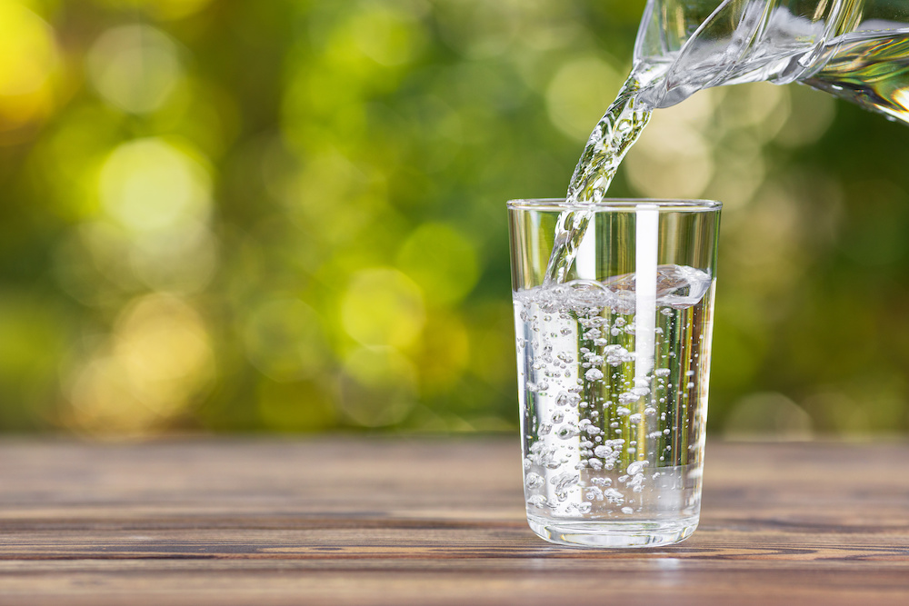 A jug pours fresh, clean water into a clear glass in front of a blurred green background.