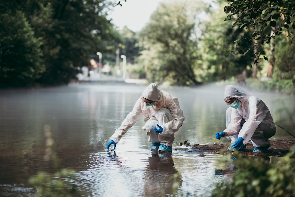 Two scientists in protective suits, gloves, and masks take water samples from a shallow river.