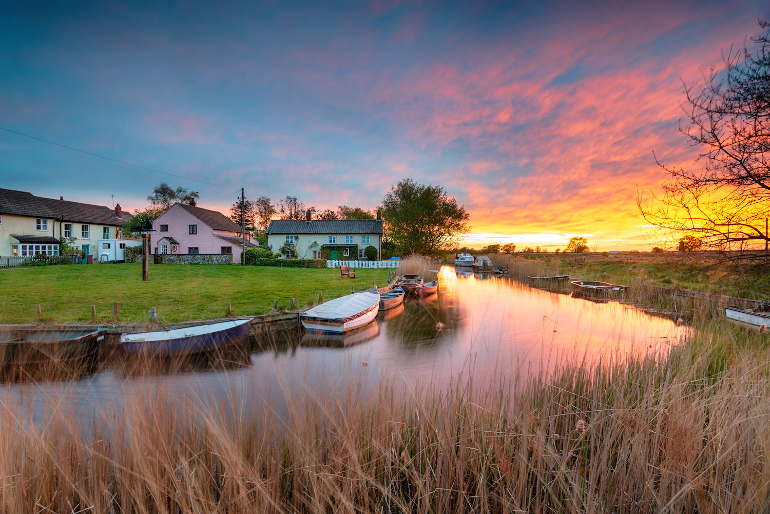 The sun sets in hues of pink and orange over a river running through a small village, with rowboats moored to the bank.