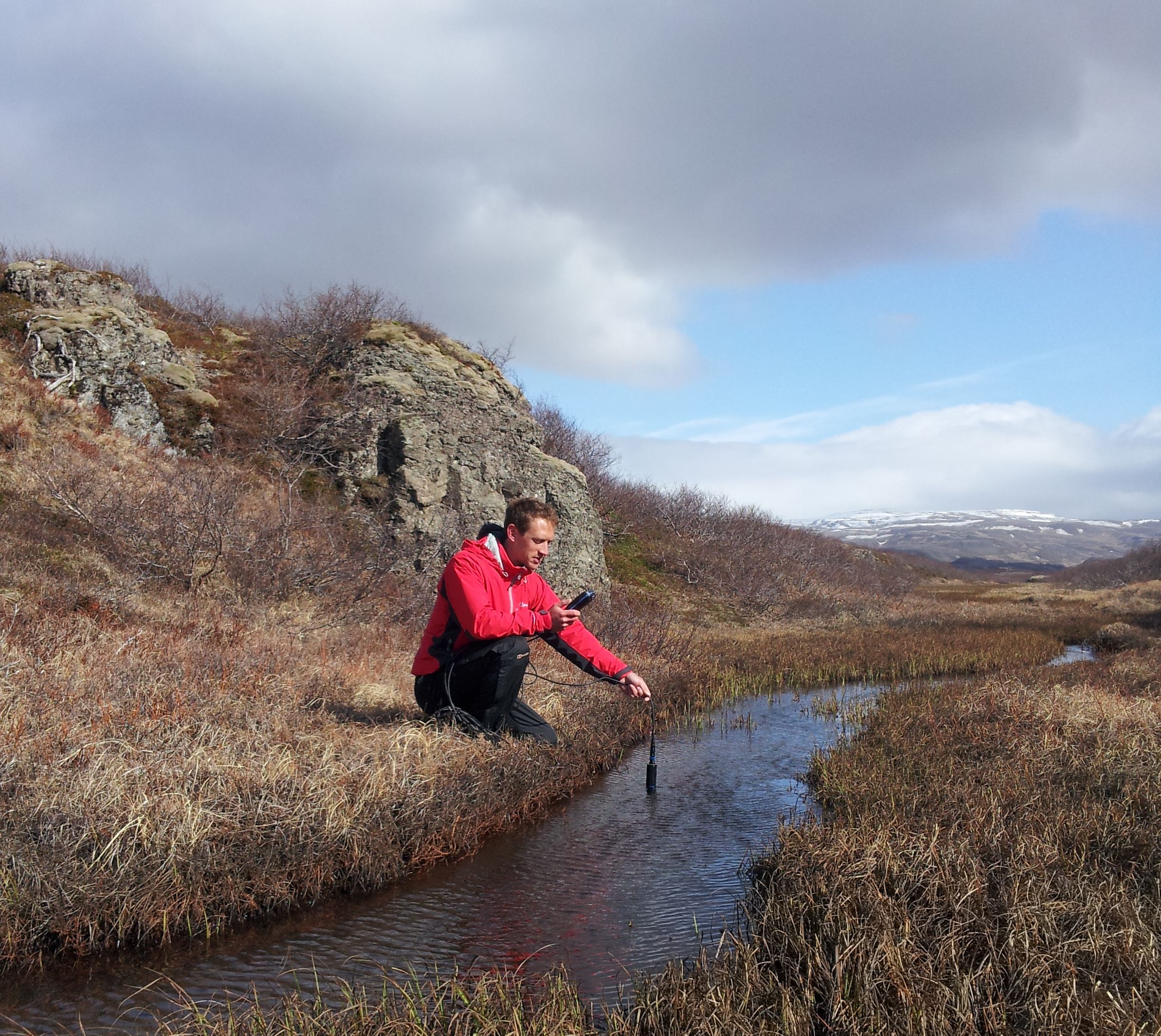 A scientist in a red coat taking a sample of water from a stream using an Aquaread monitor.